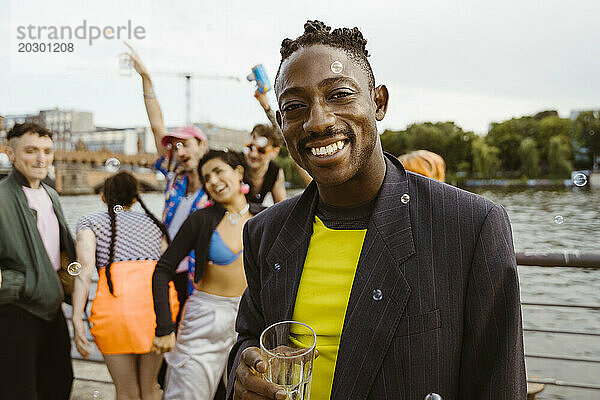 Porträt einer lächelnden nicht-binären Person  die ein Trinkglas hält und Freunde im Hintergrund genießt
