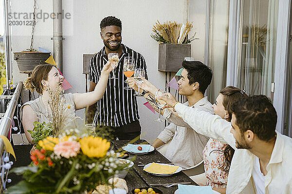 Lächelnder junger Mann  der während der Dinnerparty auf dem Balkon mit einer Gruppe männlicher und weiblicher Freunde auf Getränke anstößt