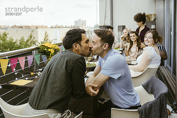 Zärtliches schwules Paar küsst sich  während es mit Freunden bei einer Dinnerparty auf dem Balkon sitzt