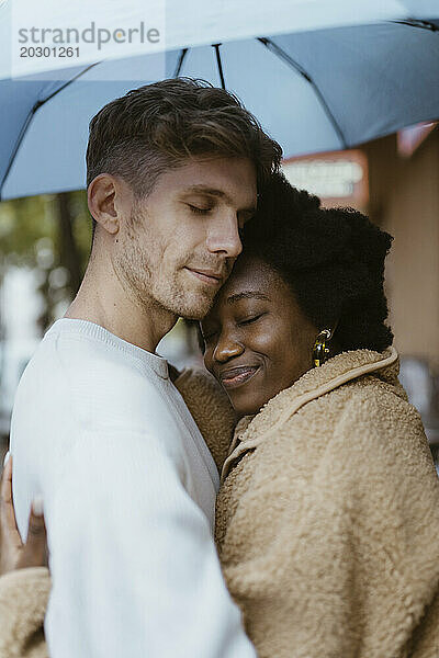 Seitenansicht eines gemischtrassigen romantischen Paares  das sich mit geschlossenen Augen unter einem Regenschirm umarmt