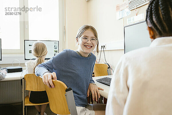 Porträt einer glücklichen Studentin  die mit Freunden im Computerunterricht in der Schule auf einem Stuhl sitzt