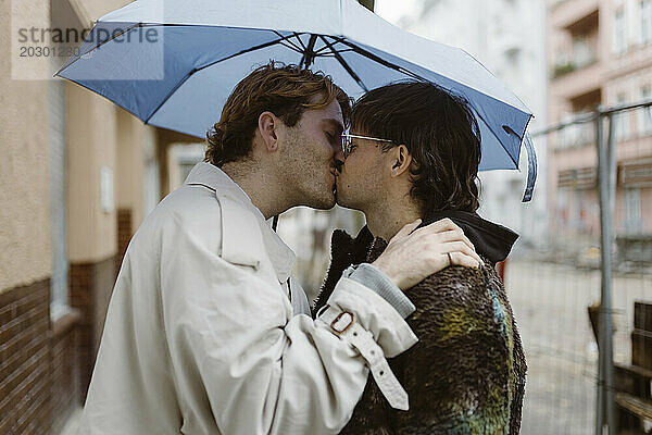Seitenansicht eines liebevollen schwulen Paares  das sich unter einem Regenschirm auf der Straße küsst