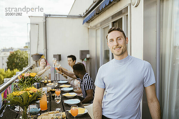 Porträt eines lächelnden Mannes mit Freunden  der während der Dinnerparty auf dem Balkon genießt