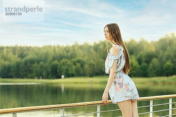 Junge Frau steht an einem Geländer und blickt auf einen ruhigen und malerischen See  der von üppigem Grün umgeben ist und einen Moment der Ruhe und Besinnung verkörpert
