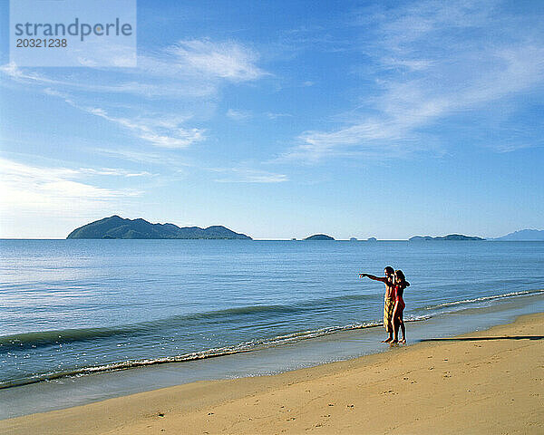 Australien. Queensland. Mission Beach. Junges Paar in Badebekleidung steht am Strand.