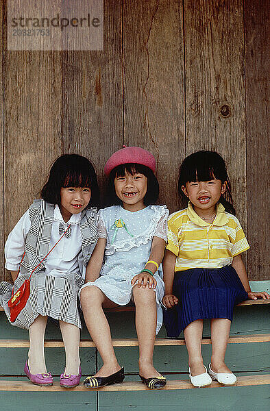 Vietnam. Da Lat. Kinder. Drei kleine Mädchen sitzen auf einer Bank in einem Holzunterstand.