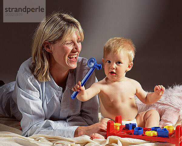 Mutter und Baby spielen drinnen mit einem Plastikspielzeughammer.