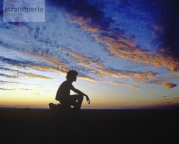 Australien. Nördliches Territorium. Silhouette eines jungen Mannes  der in der Abenddämmerung in der Wüste kauert.