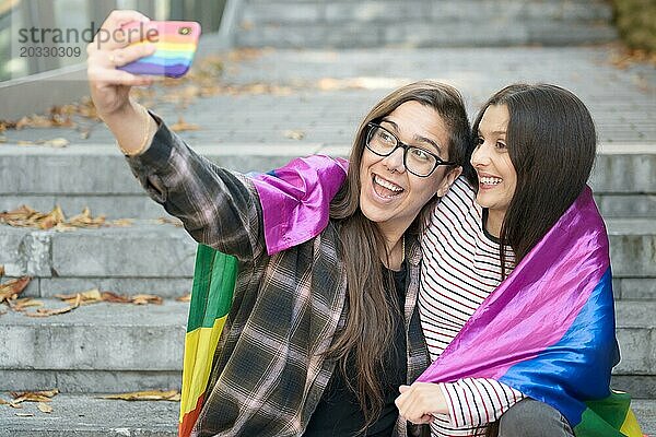 Fröhliches  glückliches lesbisches Paar  das eine Igbt Flagge hält und ein Selfie im Freien macht. Foto in hoher Qualität