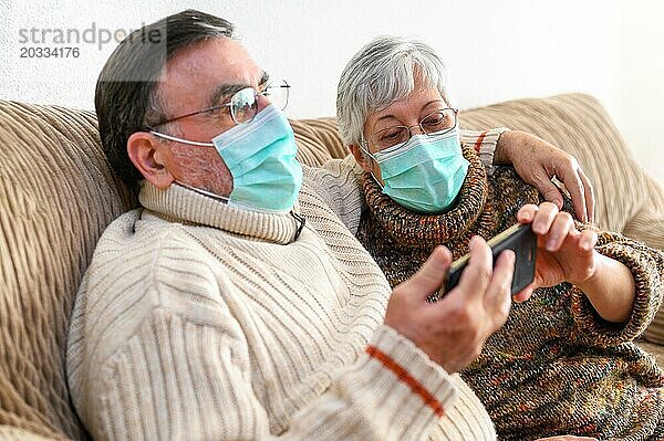 Coronavirus. Fröhliches älteres Ehepaar  das auf einem Sofa in einer Quarantänestation zu Hause sitzt und mit dem Smartphone einen Videoanruf tätigt. Älteres Ehepaar trägt Schutzmasken