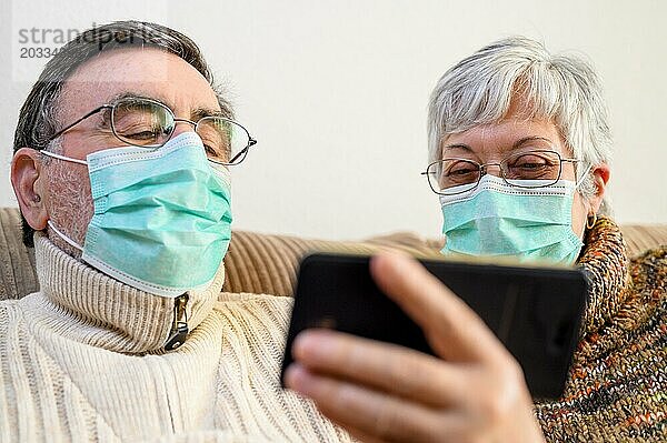 Coronavirus. Fröhliches älteres Ehepaar  das auf einem Sofa in einer Quarantänestation zu Hause sitzt und mit dem Smartphone einen Videoanruf tätigt. Älteres Ehepaar trägt Schutzmasken