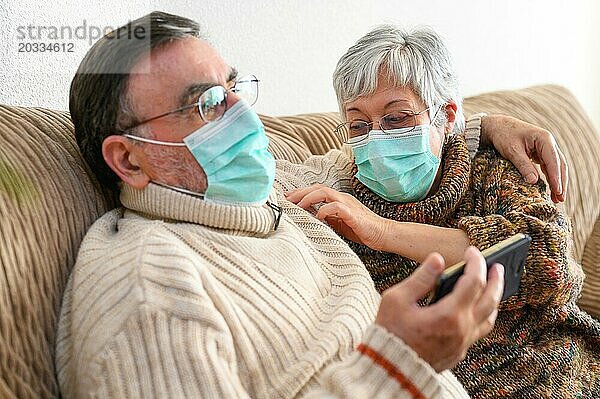 Coronavirus. Fröhliches älteres Ehepaar  das auf einem Sofa in einer Quarantänestation zu Hause sitzt und mit dem Smartphone einen Videoanruf tätigt. Älteres Ehepaar trägt Schutzmasken