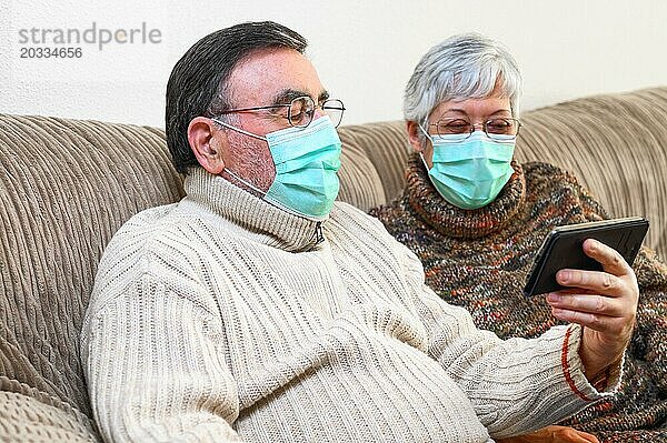 Coronavirus. Fröhliches älteres Ehepaar  das auf einem Sofa in einer Quarantänestation zu Hause sitzt und mit dem Smartphone einen Videoanruf tätigt. Älteres Ehepaar trägt Schutzmasken