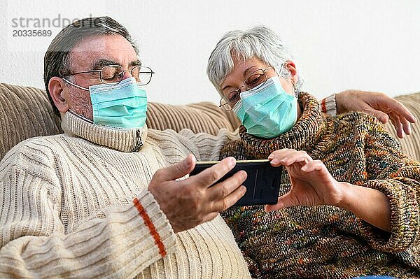 Coronavirus. Fröhliches älteres Ehepaar  das auf einem Sofa in einer Quarantänestation zu Hause sitzt und mit dem Smartphone einen Videoanruf tätigt. Älteres Ehepaar trägt Schutzmasken