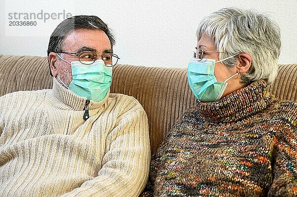 Covid 19  Lebensstil zu Hause bleiben. Fröhliches älteres Ehepaar mit Schutzmasken  das auf einem Sofa sitzt  lacht und sich gegenseitig anschaut und während der Quarantäne zu Hause positiv eingestellt ist