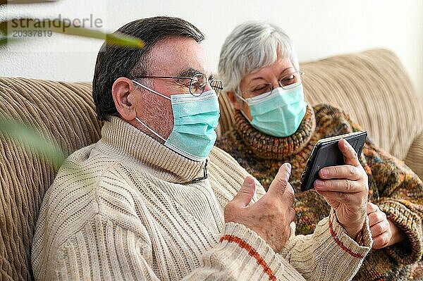 Coronavirus. Fröhliches älteres Ehepaar  das auf einem Sofa in einer Quarantänestation zu Hause sitzt und mit dem Smartphone einen Videoanruf tätigt. Älteres Ehepaar trägt Schutzmasken