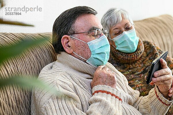 Coronavirus. Fröhliches älteres Ehepaar  das auf einem Sofa in einer Quarantänestation zu Hause sitzt und mit dem Smartphone einen Videoanruf tätigt. Älteres Ehepaar trägt Schutzmasken