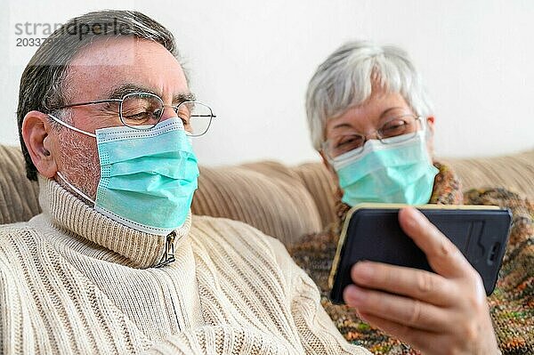 Coronavirus. Fröhliches älteres Ehepaar  das auf einem Sofa in einer Quarantänestation zu Hause sitzt und mit dem Smartphone einen Videoanruf tätigt. Älteres Ehepaar trägt Schutzmasken