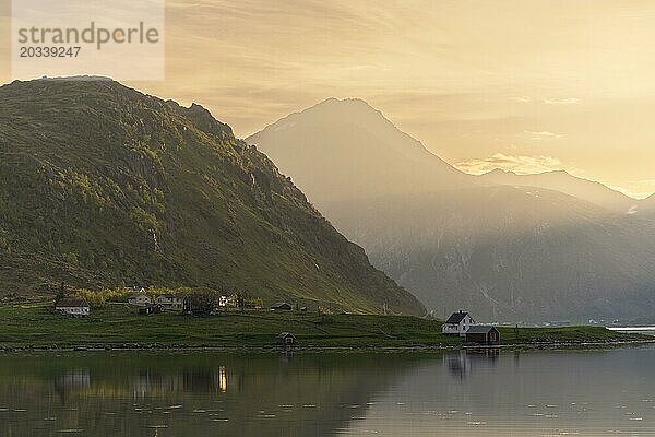 Landschaft mit Meer und Bergen auf den Lofoten. Der Fjord Flakstadpollen und einige Häuser  die zum Ort Flakstad gehören. Die Landschaft spiegelt sich im Meer. Nachts zur Zeit der Mitternachtssonne bei gutem Wetter  die Sonne scheint seitlich herein. Flakstadoya  Lofoten  Norwegen  Europa