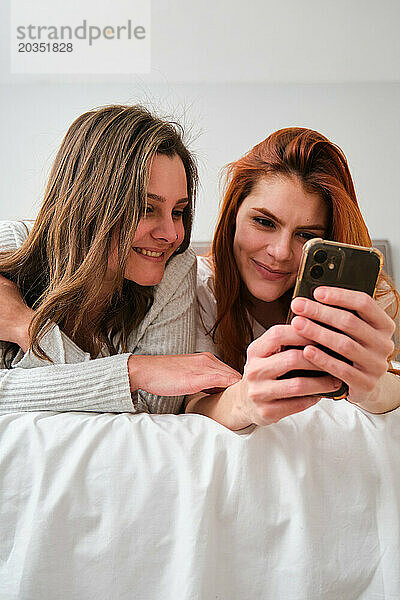 Lesbisches Paar lächelt und benutzt das Smartphone im Bett.