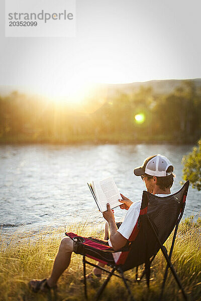 Ein sportlicher Mann liest bei Sonnenuntergang ein Buch  während er auf einer Raftingtour in Montana an einem Fluss campt.