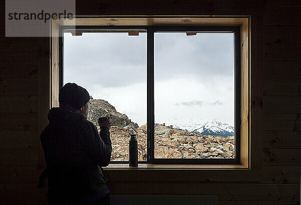 Silhouette einer jungen Frau  die Tee vor dem Fenster mit Blick auf die Berge genießt  Whistler  British Columbia  Kanada