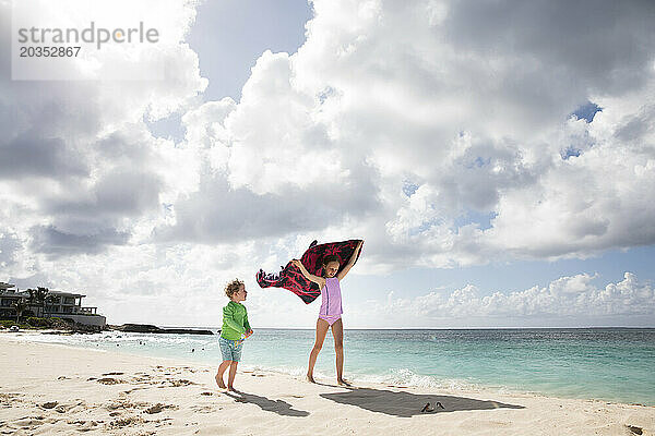 Mädchen und Bruder spielen zusammen unter einem gestreiften Handtuch am Cloudy Beach