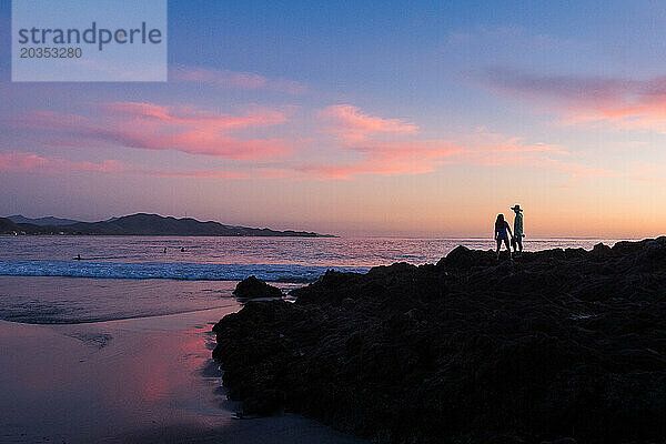 Zwei Menschen stehen bei Sonnenuntergang auf Felsen am Strand.