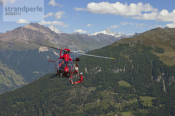 Ein Notarzt der Bergrettung hängt unter einem Hubschrauber hoch über dem Boden. Ein verletzter Wanderer wird hochgezogen. Die Schweizer Alpen im Hintergrund.