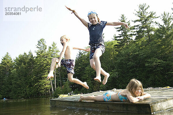 Zwei Kinder springen von einem Holzfloß in Center Harbor  New Hampshire  über einen Thrid in einen See.