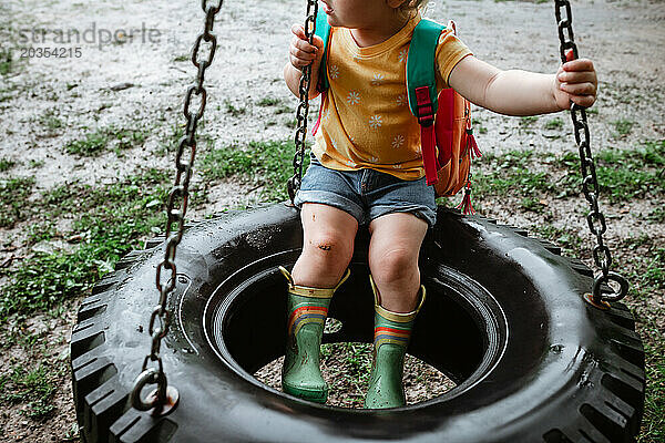 Bunt gekleidetes Kind sitzt an einem regnerischen Tag auf einer Reifenschaukel