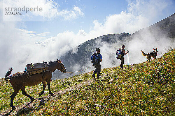 Ein Schäferhund und ein Pferd begleiten zwei Männer auf einem Bergpfad. Wandern auf dem Sentiero delle Orobie in den italienischen Alpen