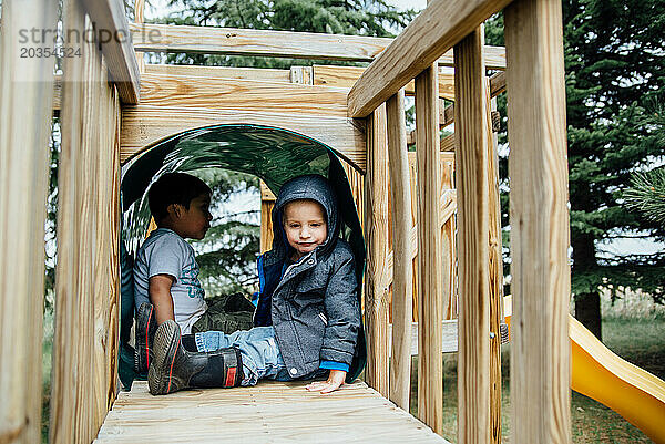 Seitenansicht von zwei kleinen Jungen  die im Tunnel einer Spielplatzschaukel sitzen.