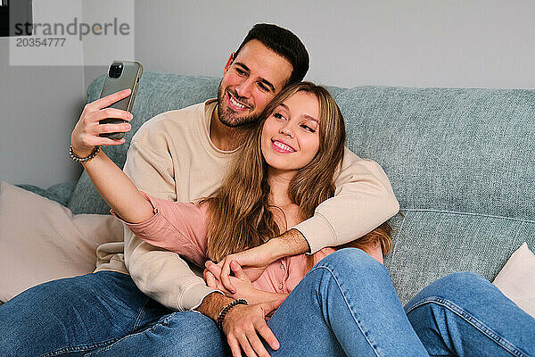 Spanisches glückliches Paar macht gemeinsam ein Selfie auf einem Sofa zu Hause.