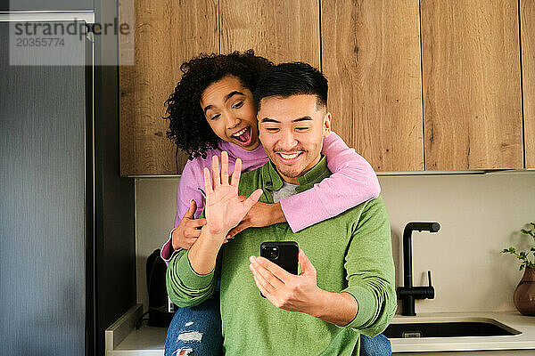 Multiethnisches glückliches Paar winkt bei einem Videoanruf in der Küche.