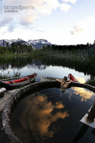 Eine Frau genießt den Sonnenuntergang im Whirlpool am Bergsee in Patagonien  Chile  während sich Wolken im Wasser spiegeln.