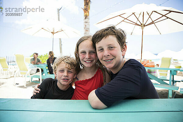 Drei Geschwister sitzen am bunten Picknicktisch und lächeln in die Kamera