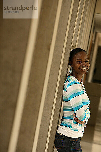 Eine junge schwarze Studentin in einer bunten Bluse lehnt lächelnd an den Säulen des Campus.