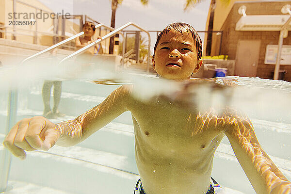 Ein kleiner Junge schwimmt mit erhobenem Kopf auf dem Wasser eines Schwimmbeckens. Auf der Schwimmbeckenleiter steht ein anderer Junge  dessen Unterkörper sich im Wasser befindet.