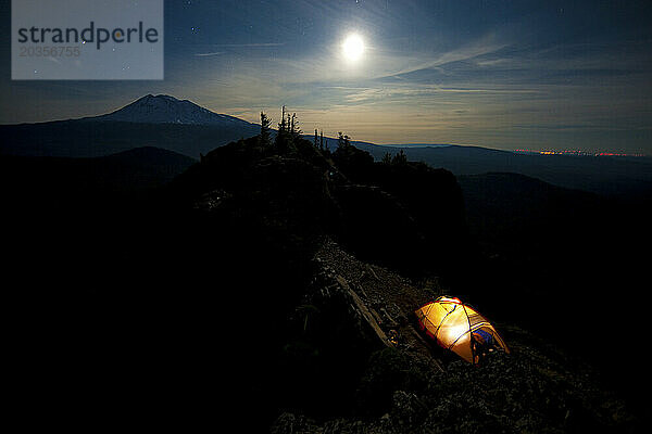 Ein Campingzelt leuchtet im Schatten felsiger Hügel unter einem hell scheinenden Mond während eines Campingausflugs entlang der Dornröschenküste im pazifischen Nordwesten