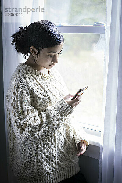 Biraciales Teenager-Mädchen schaut am Fenster auf ihr Handy