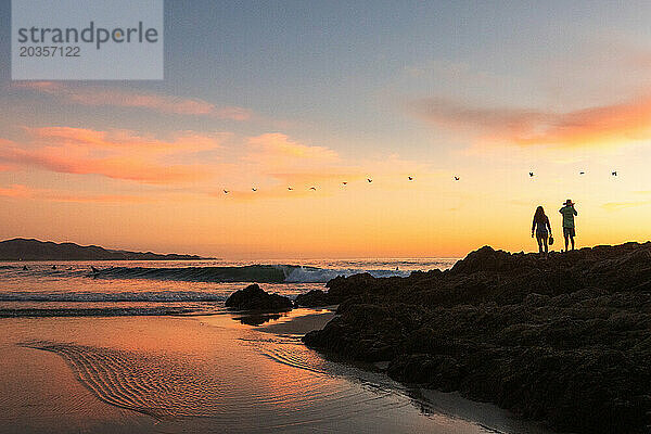 Zwei Menschen stehen bei Sonnenuntergang auf Felsen am Strand.