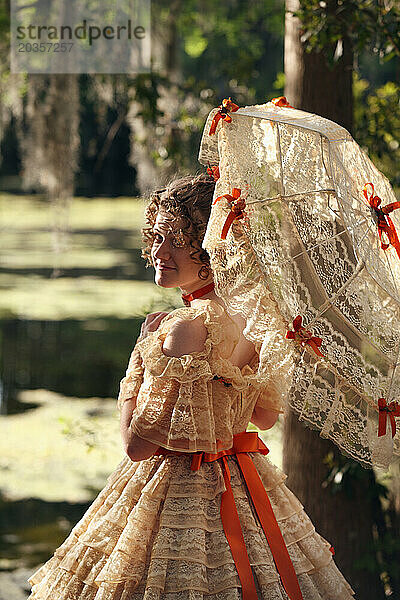 Eine junge Frau posiert in einem Southern Belle-Outfit  einschließlich eines großen  verzierten historischen Kleides und Sonnenschirms  in der Nähe eines Sees.