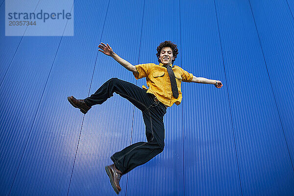 Fröhlicher junger Mann in einem gelben Hemd  der auf blauem Hintergrund springt