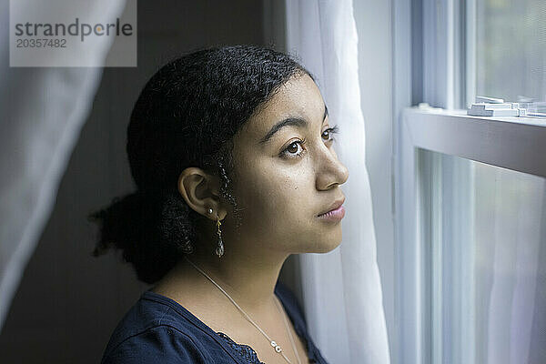 Ein gemischtrassiges Teenager-Mädchen schaut mit nachdenklichem Gesichtsausdruck aus dem Fenster