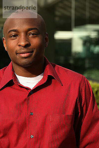 Portrait of a young African American male wearing a red shirt with a blurry modern building in the background.