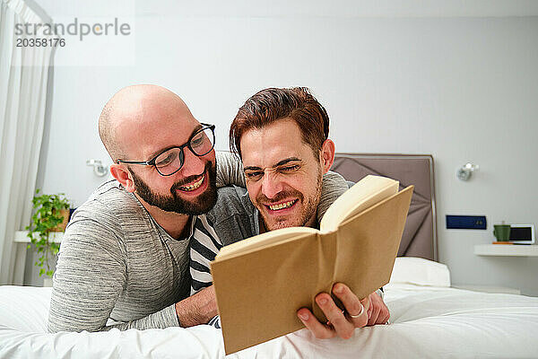 Homosexuelles Paar liest ein Buch  umarmt sich und lacht gemeinsam im Bett.