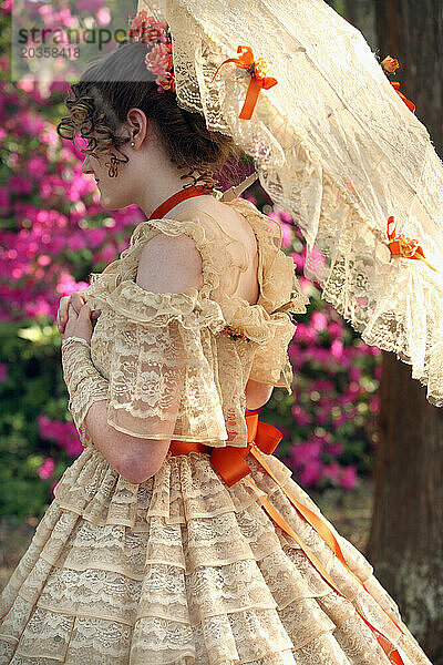 Eine junge Frau posiert in einem Southern Belle-Outfit  einschließlich eines großen  verzierten historischen Kleides und eines Sonnenschirms  unter einem Baum.