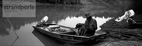 Panorama eines afroamerikanischen Mannes  der in seinem Boot sitzt und seinen Hund auf dem Mississippi umarmt.