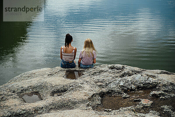 Weite Rückansicht von zwei Teenager-Mädchen  die auf Felsen in der Nähe eines Sees sitzen.